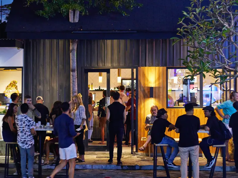 The bustling evening scene outside Tygr Sushi restaurant in Canggu, with diners enjoying food and drinks at outdoor tables under the warm glow of the modern, industrial-style building.