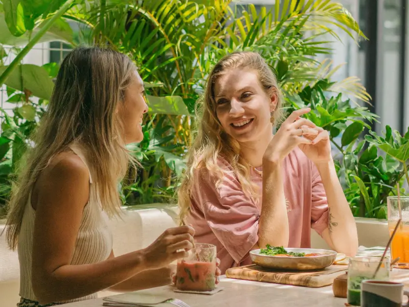 Two smiling women enjoying a healthy meal and drinks at Lush Kitchen, a bright cafe surrounded by lush green plants, creating a vibrant and tropical dining atmosphere.