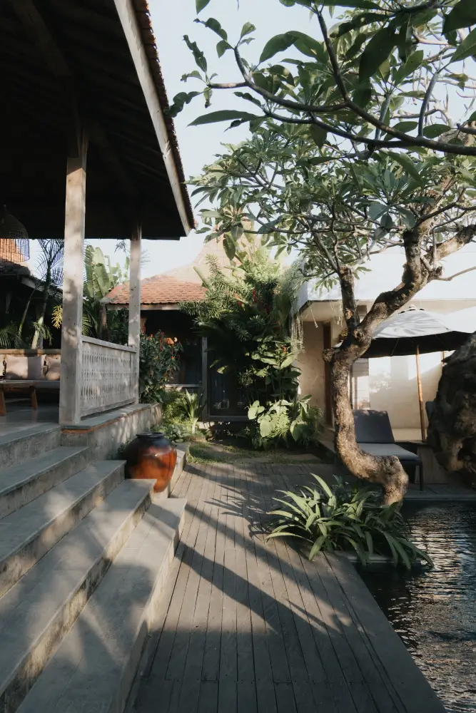 The comfortable terrace of a unit at Rimbun Villas Canggu, with concrete steps leading from a wooden deck that runs alongside a shared swimming pool, surrounded by lush garden landscaping and a frangani tree.