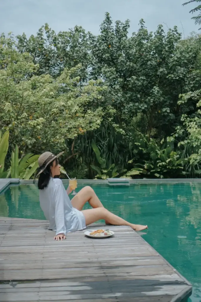 A woman in a sun hat and white shirt relaxing on the wooden deck of the Rimbun Hotel pool, holding a glass of juice and enjoying a light snack with her feet near the water.