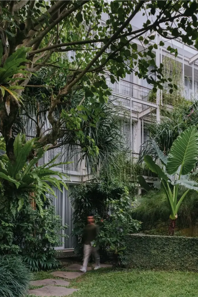 A guest with motion blur walks along a stone path in the Rimbun Hotel garden, surrounded by an abundance of lush green plants and trees that grow alongside the modern facade of the hotel.