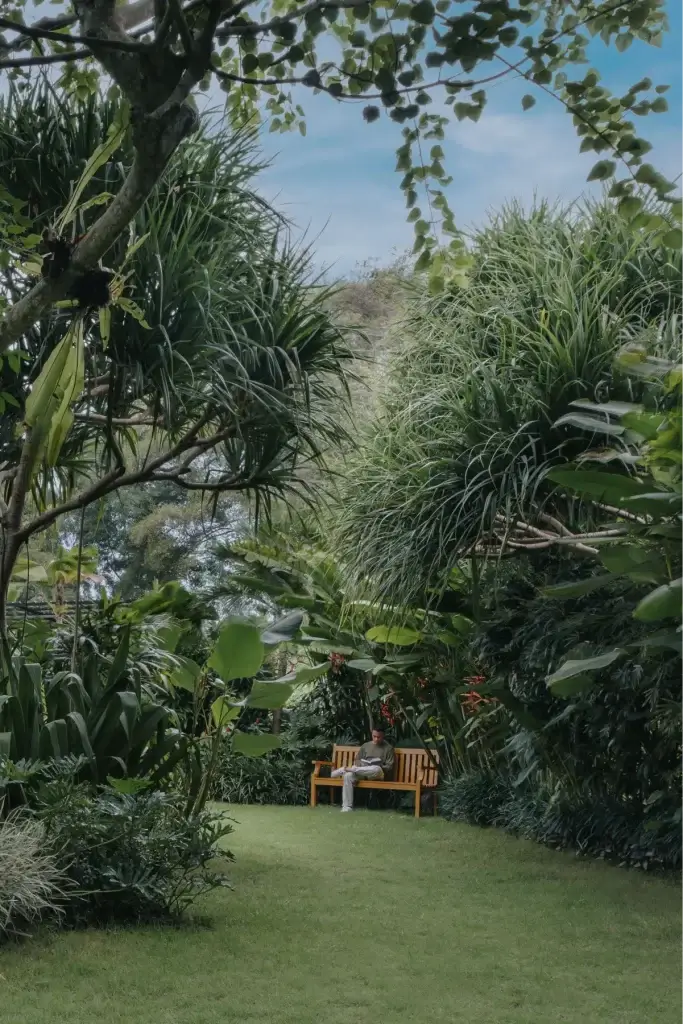 A guest sits on a wooden bench in a secluded part of the Rimbun Hotel garden, completely surrounded by towering, dense green tropical plants and trees, creating a private, tranquil nook.