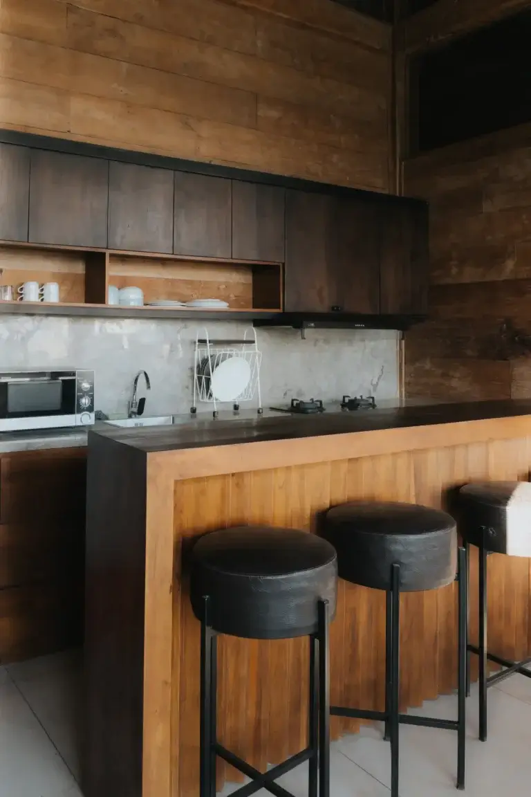 The stylish and functional mini pantry at Rimbun Canggu Villa, featuring a wooden breakfast bar with black leather stools, dark wood cabinetry, a concrete backsplash, a gas stove, and a microwave.