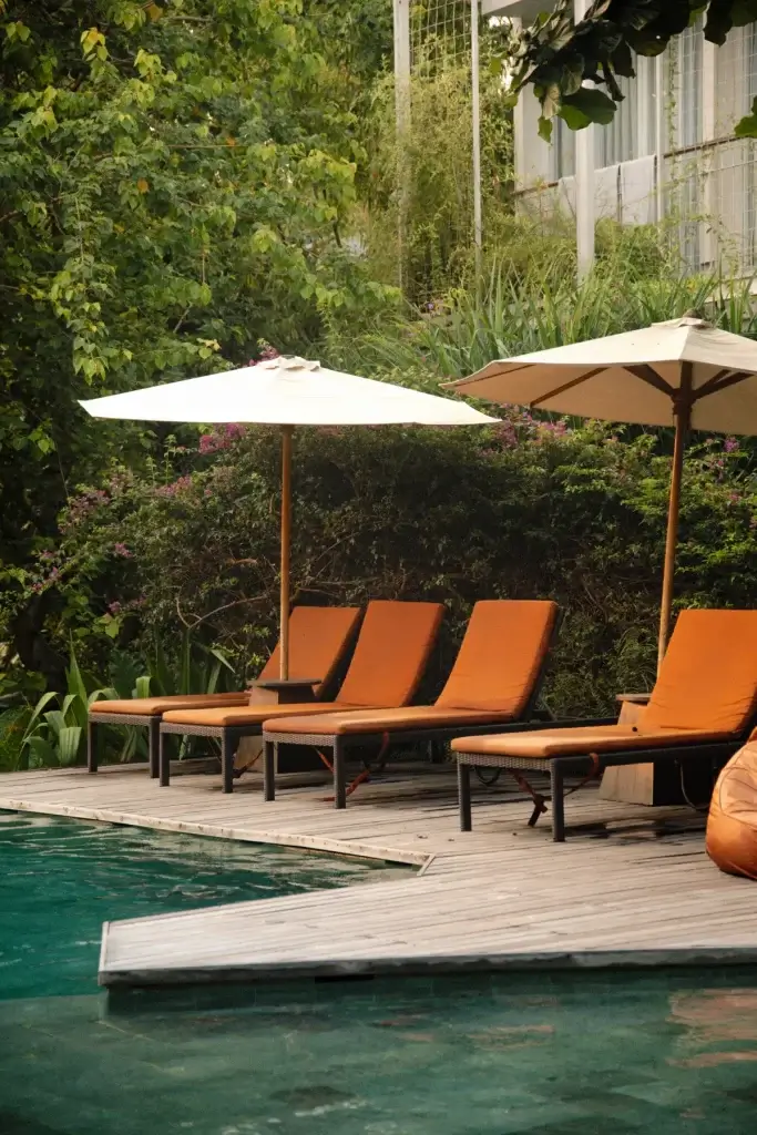 A row of comfortable orange sun loungers with large beige umbrellas set up on the wooden deck next to the swimming pool at Rimbun Hotel, surrounded by a lush green garden.