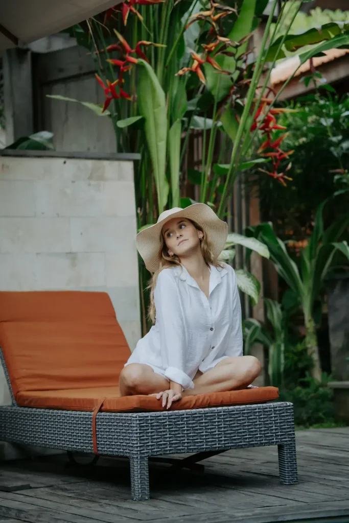 A woman wearing a white shirt and a wide-brimmed sun hat sits elegantly on a cushioned, dark-weave sun lounger at the Rimbun Villa poolside, with tall, vibrant heliconia flowers in the background.