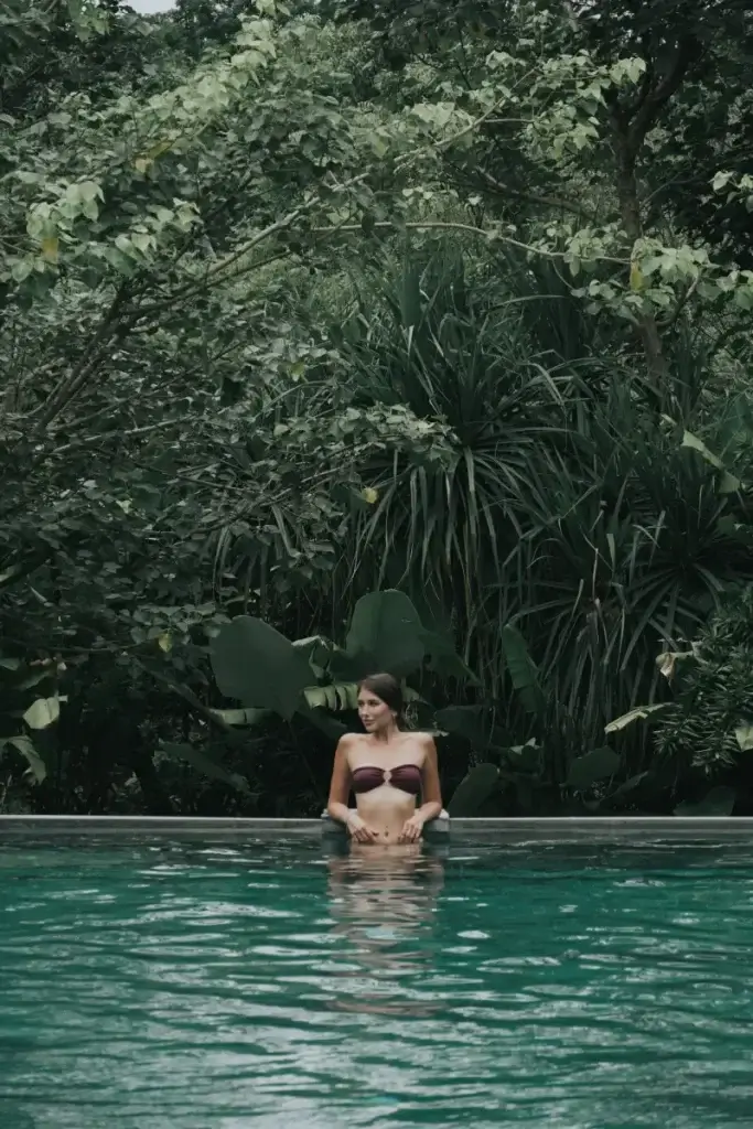 A woman in a bikini relaxing in the Rimbun Hotel swimming pool, with a stunning backdrop of dense, towering, and lush tropical jungle foliage, creating a secluded oasis feel.