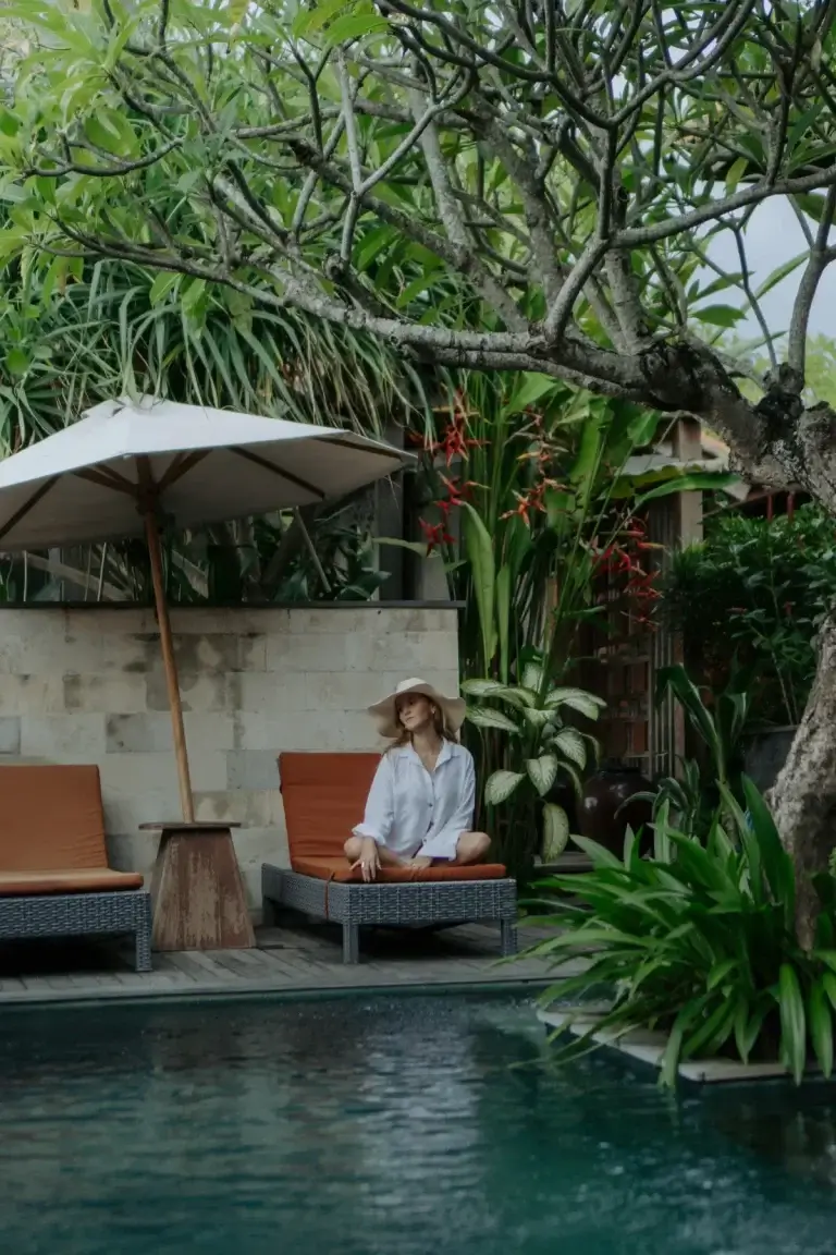 A woman in a white shirt and sun hat relaxes on a comfortable orange sun lounger by the Rimbun Villa shared pool, sitting under the shade of a large, beautiful frangipani tree and surrounded by lush tropical gardens.