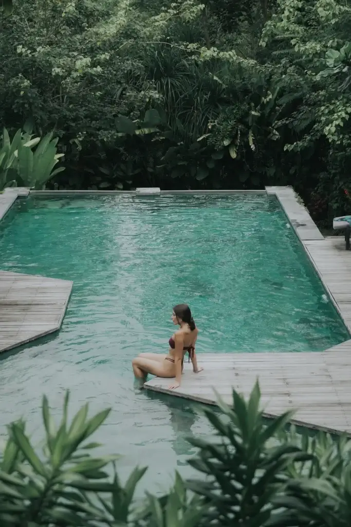 A woman in a bikini sitting on the edge of the unique wooden deck that extends into the turquoise water of the Rimbun Hotel pool, surrounded by lush greenery.