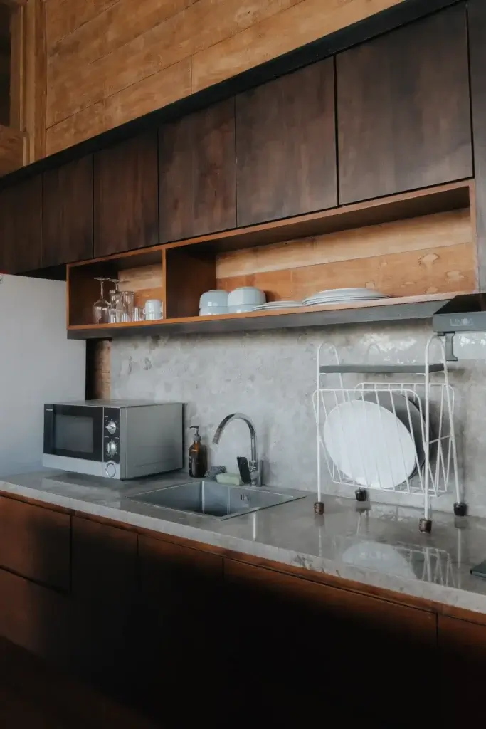 A close-up of the mini pantry's countertop area at Rimbun Villa, showing a stainless steel microwave, a sink with a modern faucet, a dish rack, and concrete countertops against a backdrop of wooden cabinets.