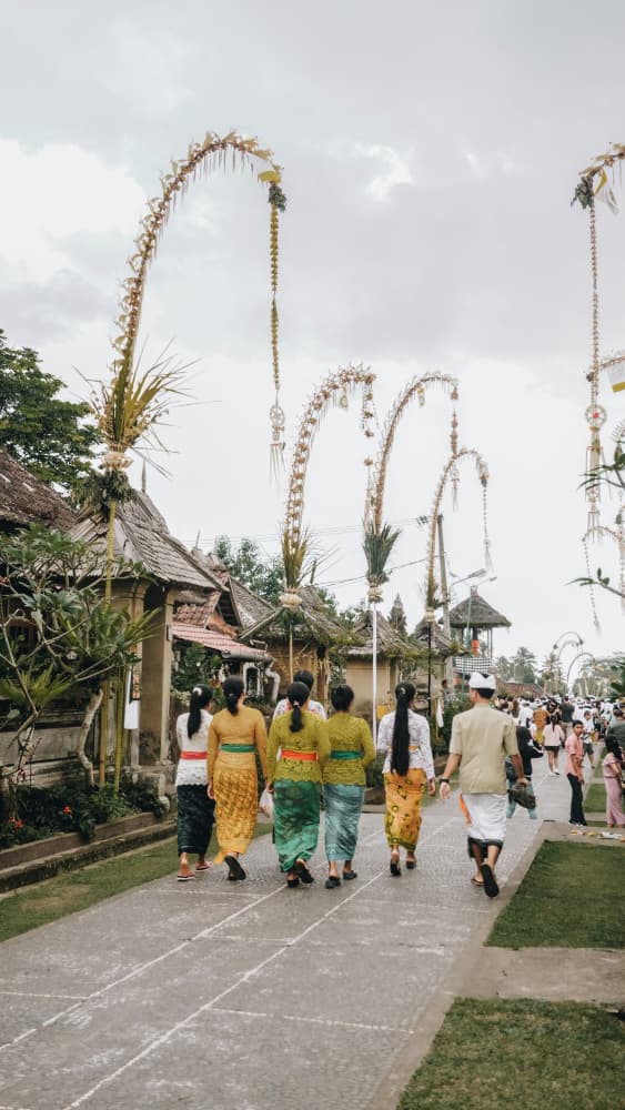 People in traditional Balinese attire walking past penjor poles during Kuningan ceremony in Penglipuran Village.