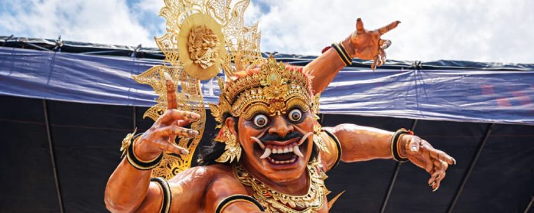 Vibrant Balinese Ogoh-ogoh statue with multiple arms and fierce expression under a blue sky.
