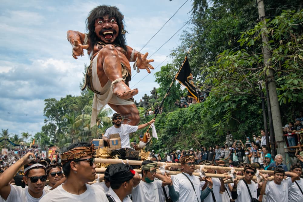 Large brown monster effigy being carried by men in white t-shirts during a Balinese parade.