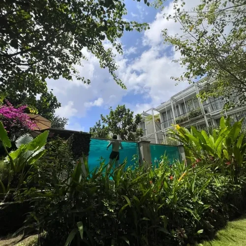 A creative shot from a guest showing a man swimming in Rimbun Canggu's unique glass-walled swimming pool, viewed from the garden with lush plants in the foreground and the hotel building behind.