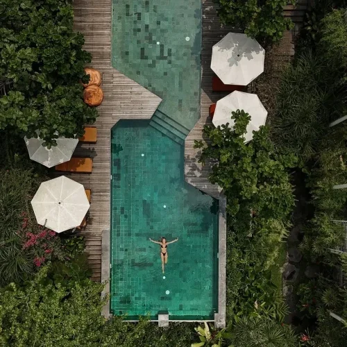 An aerial drone shot of a woman in a bikini floating peacefully in the uniquely shaped turquoise swimming pool at Rimbun Canggu, surrounded by a wooden deck, white umbrellas, and lush tropical greenery.