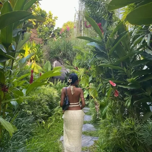 A guest's photo taken from behind a stylish woman with a flower in her hair, as she walks along a stone path through the dense, vibrant tropical gardens of Rimbun Canggu.