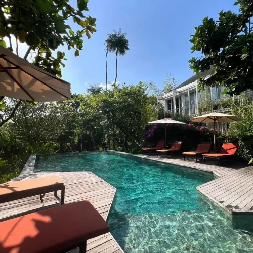 A guest's perspective of the Rimbun Canggu pool from a wooden deck, showing the sparkling turquoise water, orange lounge chairs, and beige umbrellas set against a backdrop of lush trees and the hotel building.